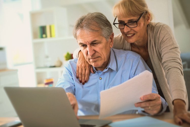 Ältere Mann sitzt mit Unterlagen in der Hand vor einem Computer. Hinter Ihm steht eine ältere Frau und legt die Hand auf seine Schulter. Beide schauen auf den Computer.
